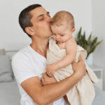 indoor-shot-brunette-man-loving-father-wearing-white-casual-t-shirt-holding-his-infant-daughter-wrapped-towel-kissing-her-head-with-love-care-posing-home-light-room-scaled.jpg